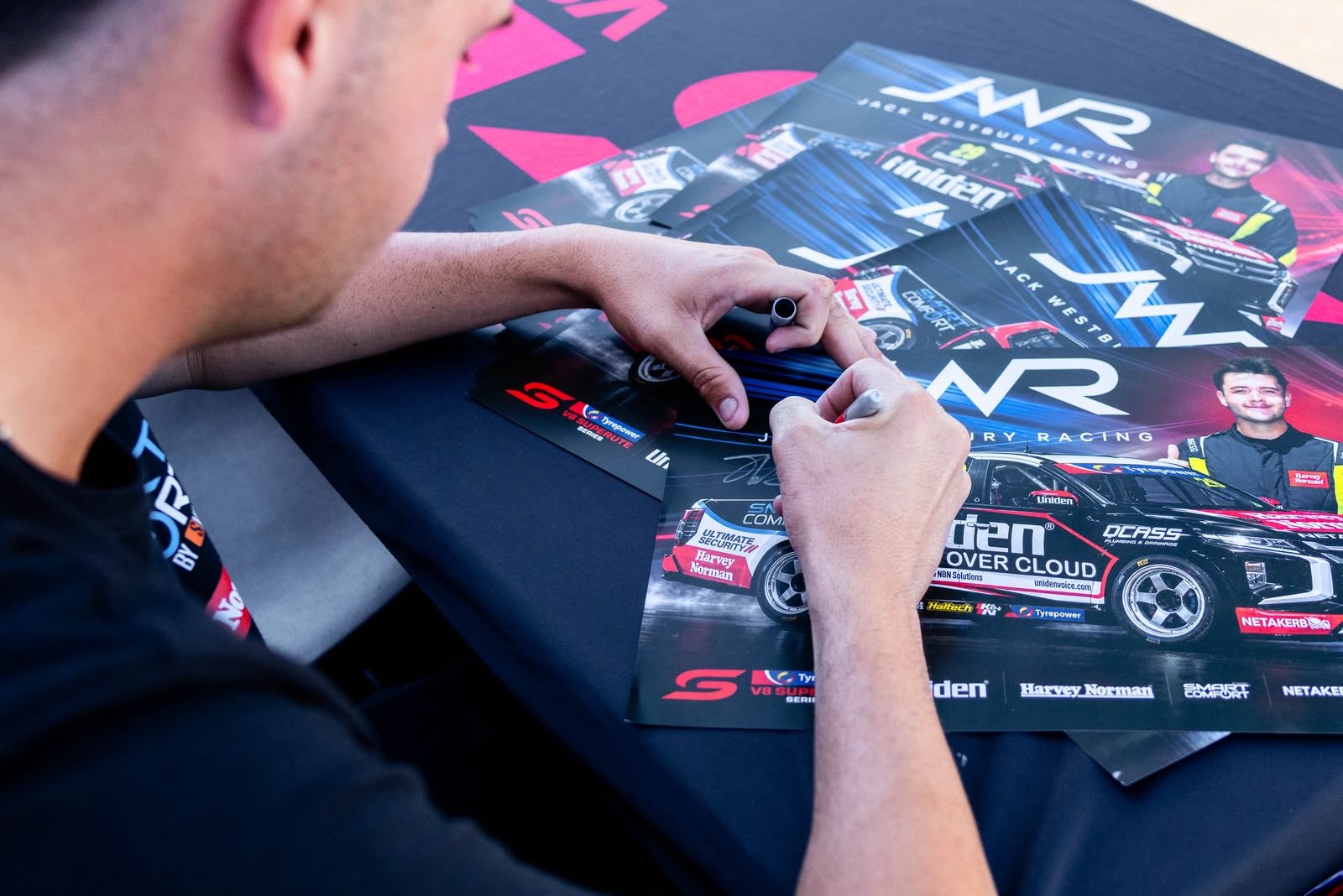 Close-up of Jack Westbury signing Jack Westbury Racing posters featuring the #29 Uniden Voice Over Cloud Ute
