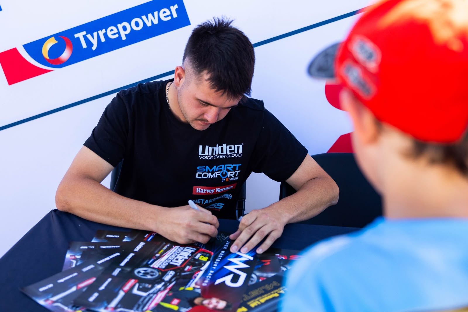 Jack Westbury in a Uniden Voice Over Cloud t-shirt signing posters for a young fan at the autograph session