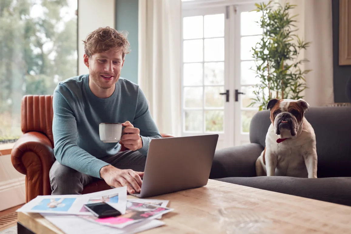 A sole trader working from home on a laptop with a coffee and the family dog beside him